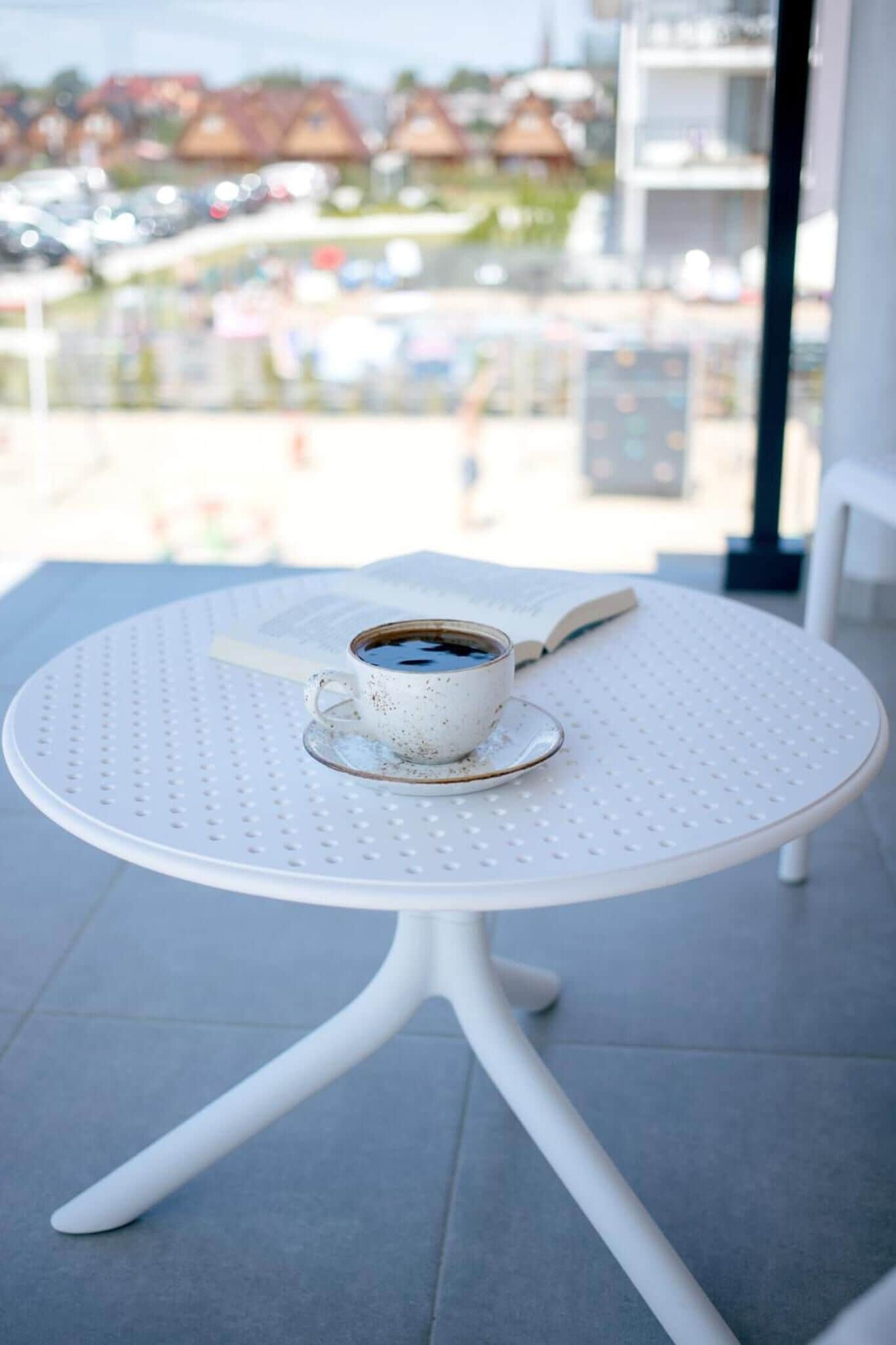 A white metal table with a cup of coffee and a book on it, set on a balcony with a view of buildings and a parking lot.