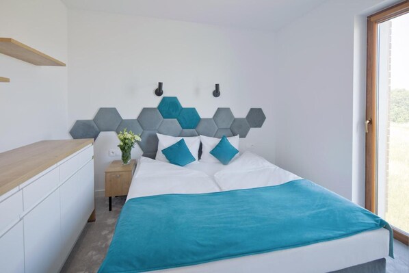 Un dormitorio con cama, una mesita de noche con un florero, un estante de madera sobre la cama y una ventana con vistas a la vegetación.