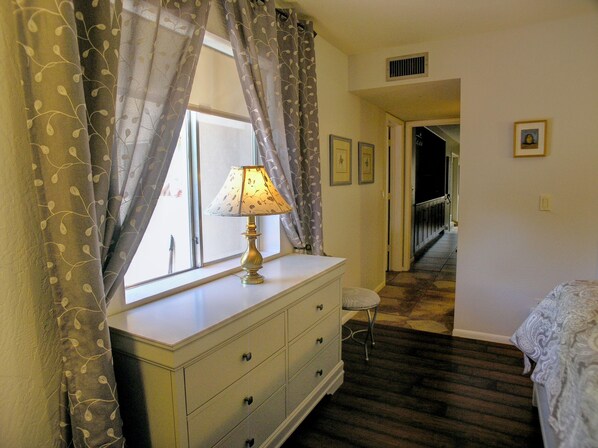 A bedroom with a dresser, a lamp, a bed, a window with curtains, a chair, and a hallway leading to another room. - Sedona Red Rock Retreat | Hot Tub & RV Parking Near Chapel (Sedona)