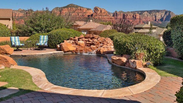 Uma piscina com cascata, rodeada por um terraço de tijolos e duas espreguiçadeiras.