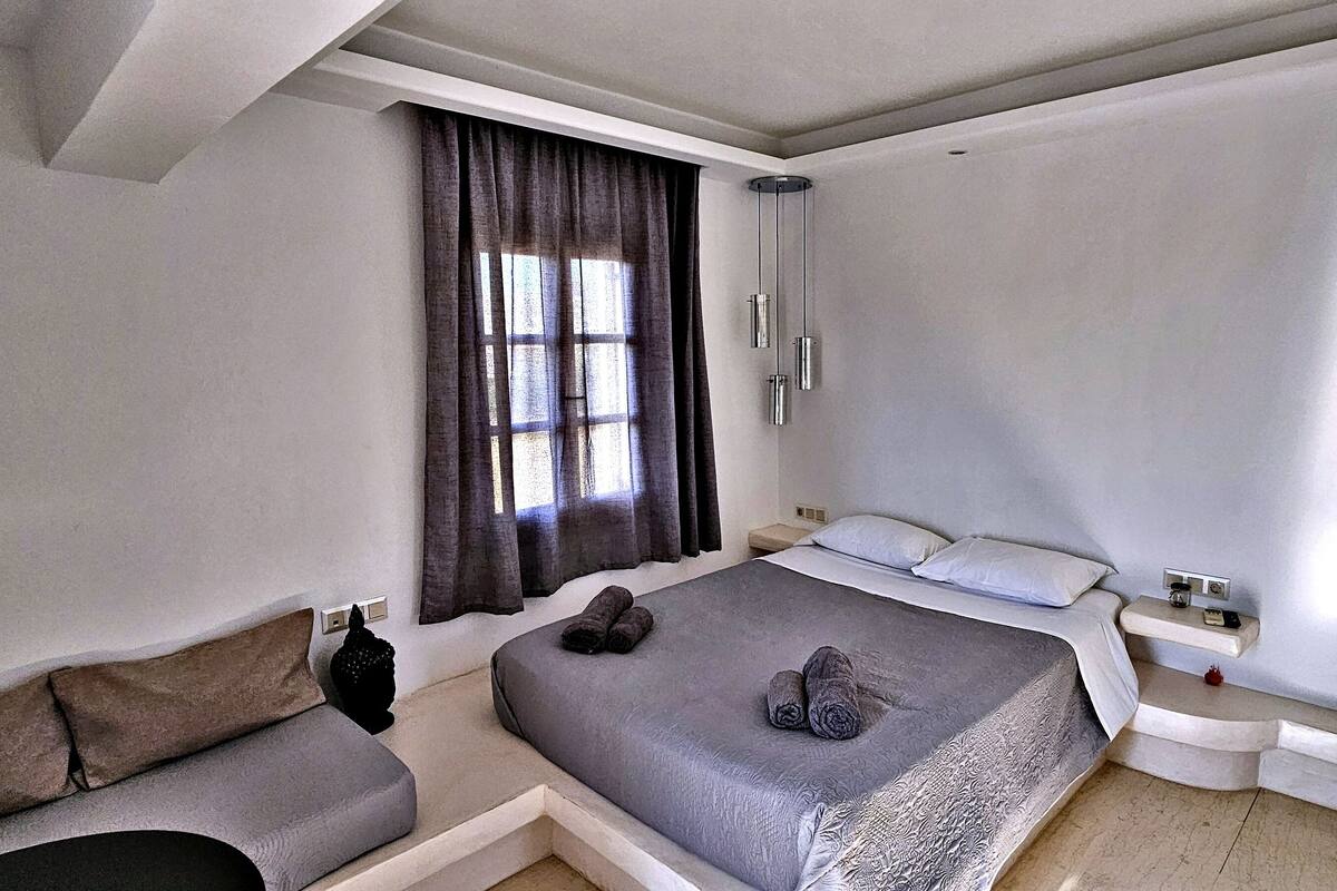 Un dormitorio moderno con una cama grande, una mesita de noche, una repisa, una ventana con cortinas y un banco empotrado.