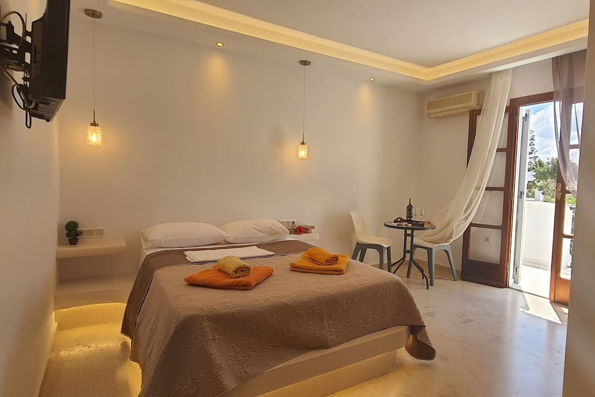 Habitación de hotel con cama, una pequeña mesa con sillas, televisor montado en la pared y ventana con cortinas.