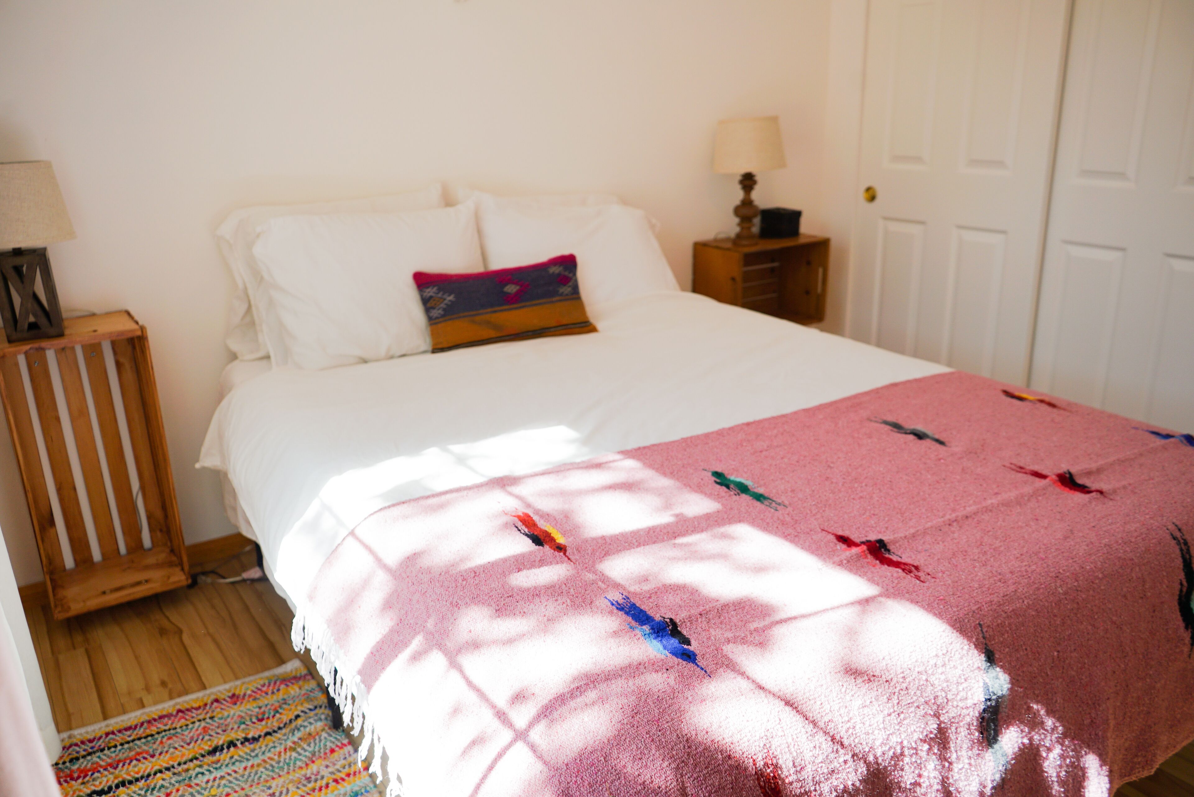 Un dormitorio con cama, almohadas, manta, mesita de noche, lámpara y una puerta.
