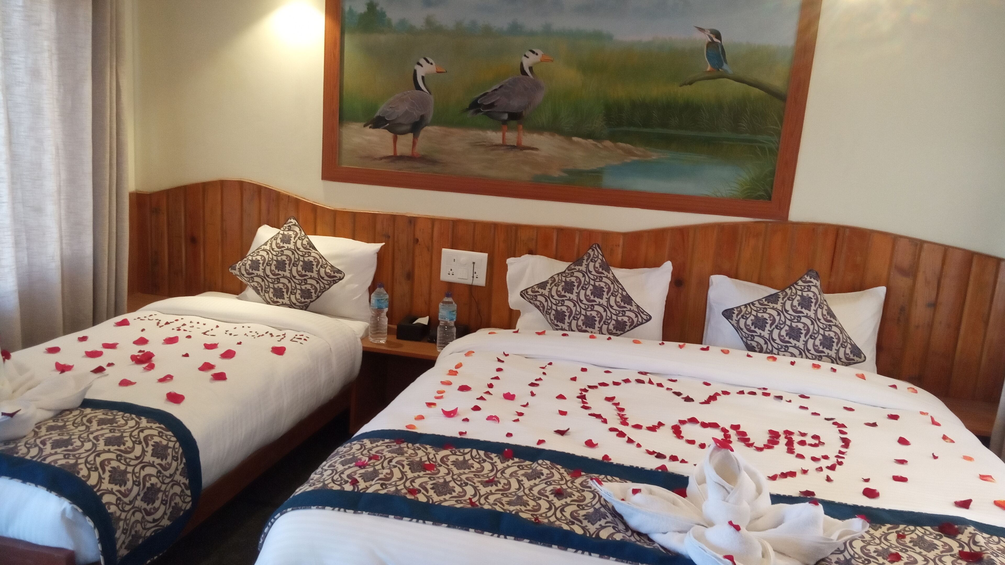 A hotel room with two beds, each with a heart-shaped arrangement of rose petals and a white towel. There is a painting of birds on the wall above the beds.