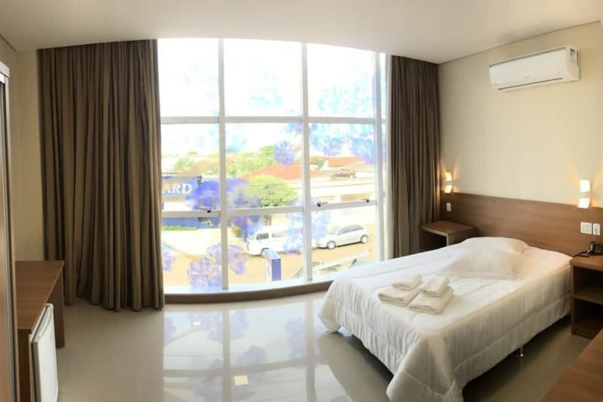 Een hotelkamer met een groot raam, een bed met witte lakens, een nachtkastje en een bedtafel.