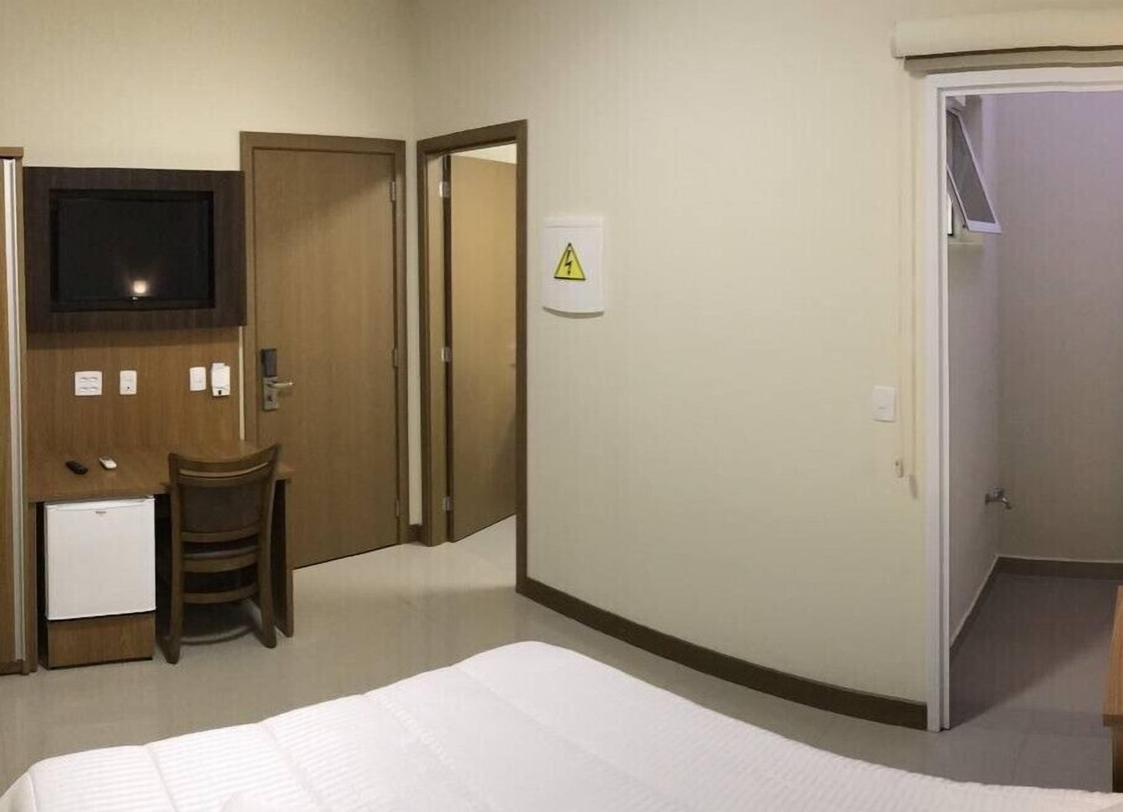 Une chambre d’hôtel comprenant un lit, un bureau avec une chaise, une télévision et une porte sur laquelle est apposé un panneau d’avertissement.