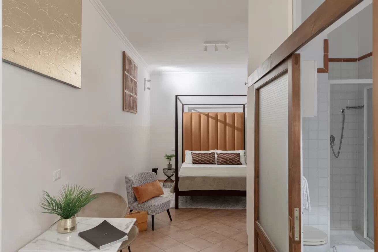 A compact hotel room with a bed, a small table with a plant, a chair, and a shower area.