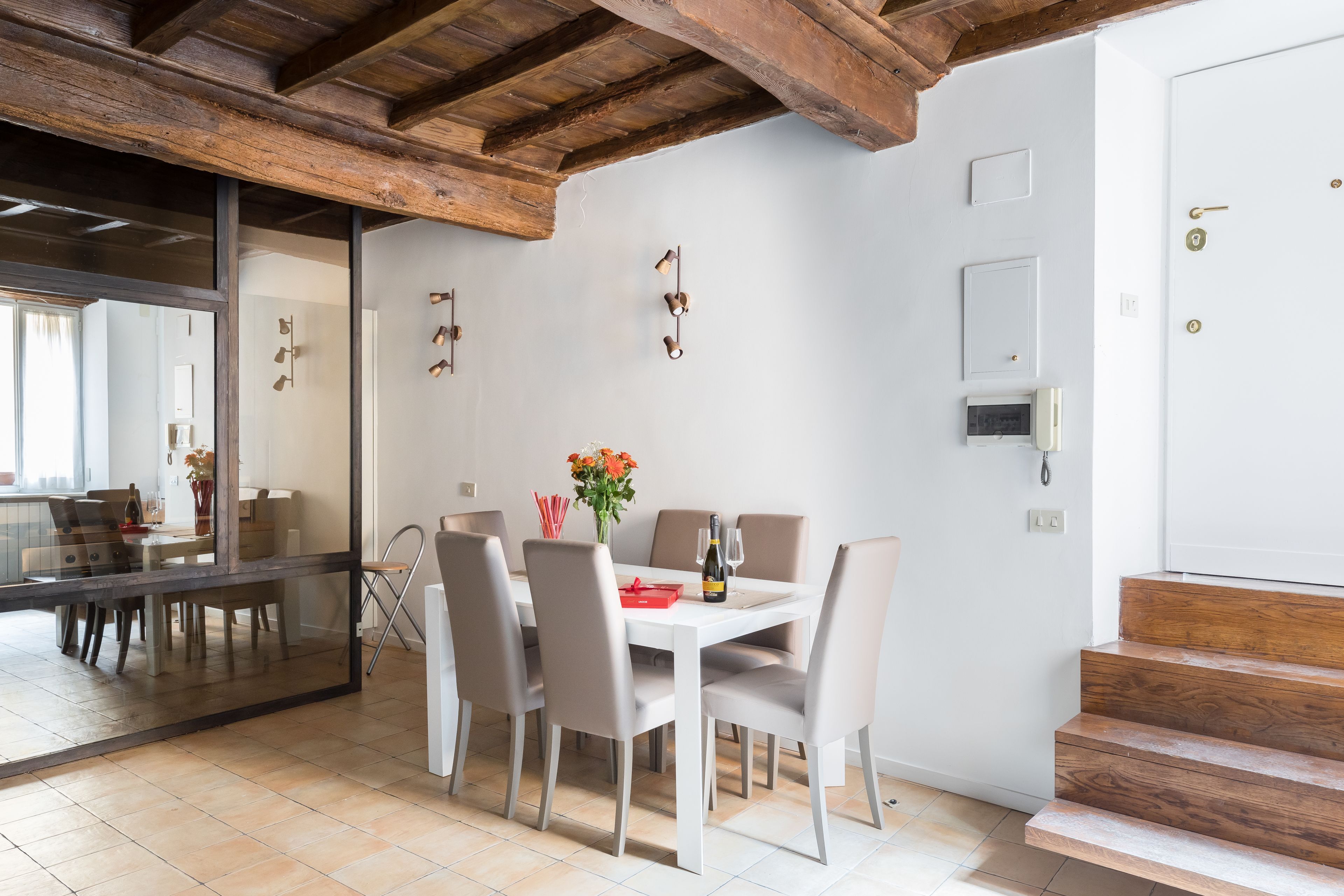 A dining area with a white table and chairs, wooden beams on the ceiling, and a view into another room.