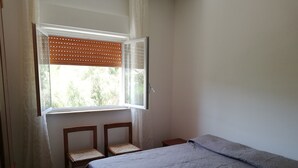 Ein kleiner, schlichter Raum mit einem Bett, zwei Stühlen, einem Fenster mit Jalousien und einem gemusterten Boden.