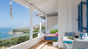 A balcony with a bed, a table with a bowl of fruit, and a view of the sea and hills. - Ferma Hill Apartments (Ierapetra)