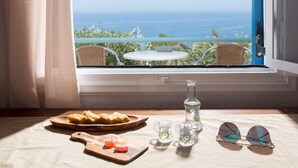 A table with a plate of food, a glass pitcher, and a pair of sunglasses by a window overlooking a view of the sea. - Ferma Hill Apartments (Ierapetra)