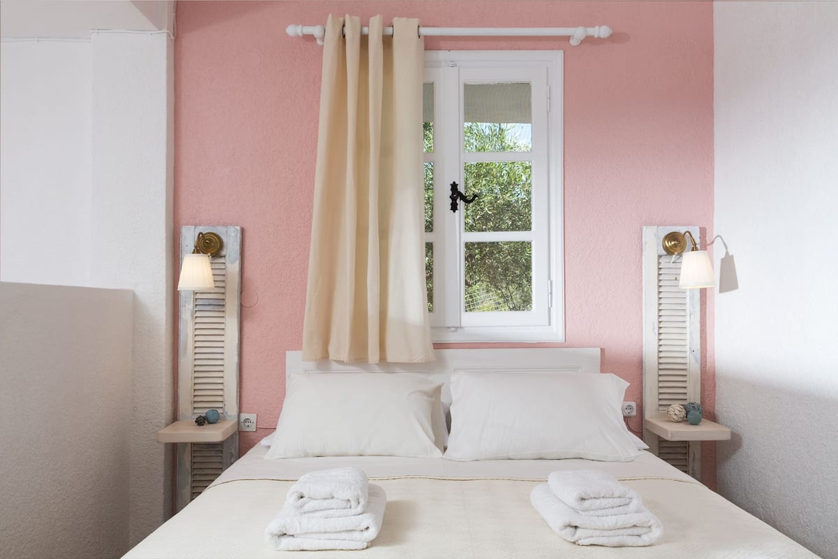 Ein Schlafzimmer mit einem Bett, zwei Nachttischen, einem Fenster mit Vorhängen und Blick ins Grüne.