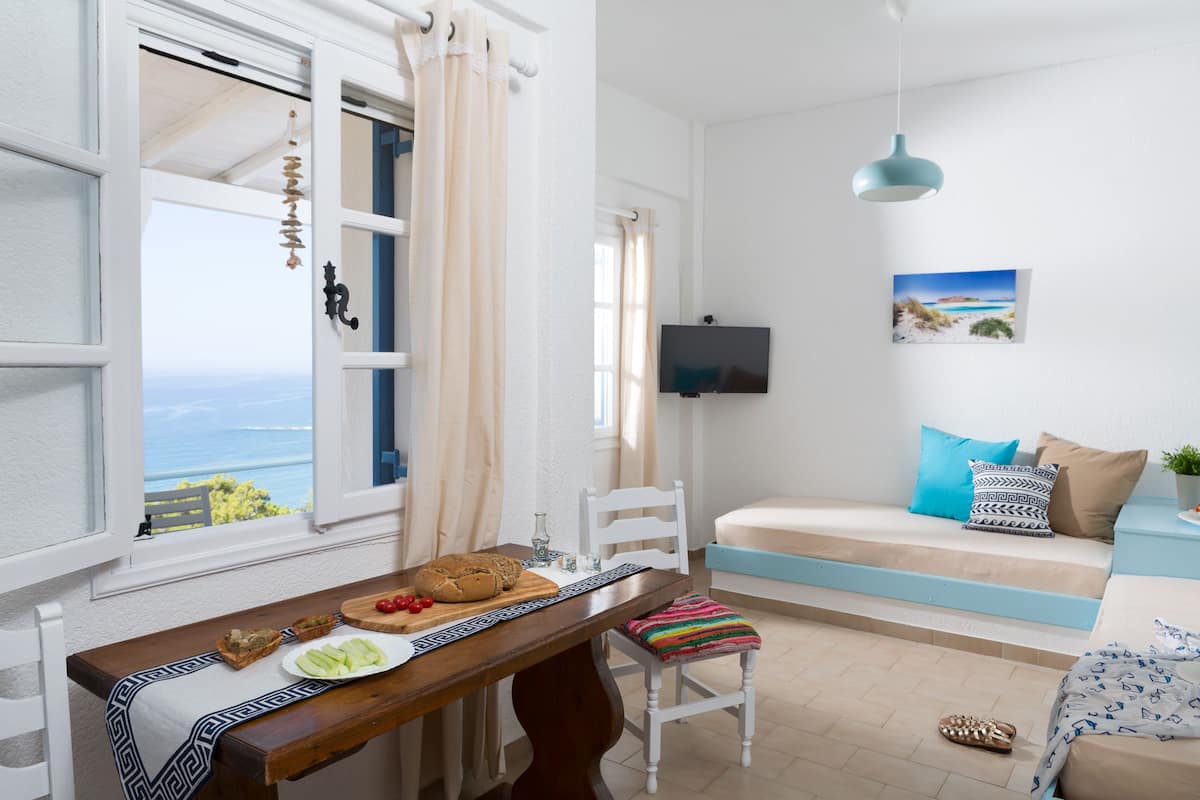 Ein gemütliches Zimmer mit Holztisch, gedeckter Einkehr, einem weißen Stuhl, einer Couch mit bunten Kissen, einem Fernseher und Meerblick durch ein Fenster.
