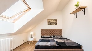 A bedroom with a sloped ceiling, a bed with a tufted headboard, a radiator, a small bedside table with a lamp, and a wall-mounted shelf with a plant. - Apart-Hostel ZERO (Bratislava)