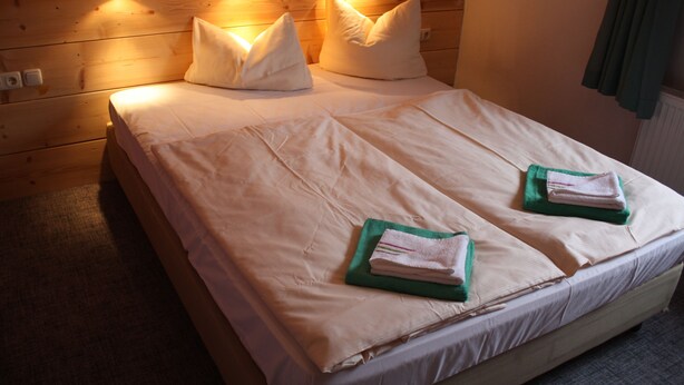 Una cama bien hecha con sábanas blancas, dos almohadas y dos toallas dobladas sobre un cabecero de madera.