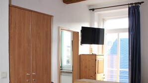 Una habitación con armario de madera, televisor montado en la pared, mesa de comedor con sillas y ventana con cortinas.