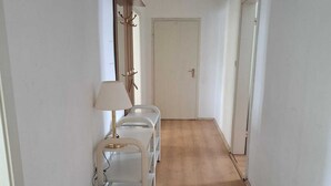 Un pasillo estrecho con suelo de madera, una puerta blanca al final y un perchero con una lámpara y un bolso a la izquierda.