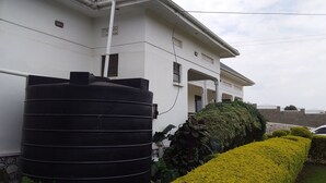 A building with a large black water tank attached to the side, a white exterior, and a well-maintained hedge. - Trust White Hotel (Bushenyi)