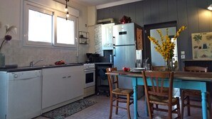 Keittiössä on valkoiset kaapit, ruostumattomasta teräksestä valmistettu jääkaappi, ruokapöytä tuoleineen sekä maljakko, jossa on keltaisia kukkia.