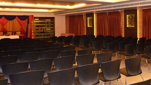 Un salón de conferencias con filas de sillas negras, un escenario con cortinas rojas y doradas, y una mesa cubierta con un mantel blanco.