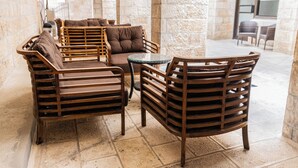 A seating area with wooden chairs and a round table, stone walls, and a view of the outside. - New Gate Hostel (Jerusalem)