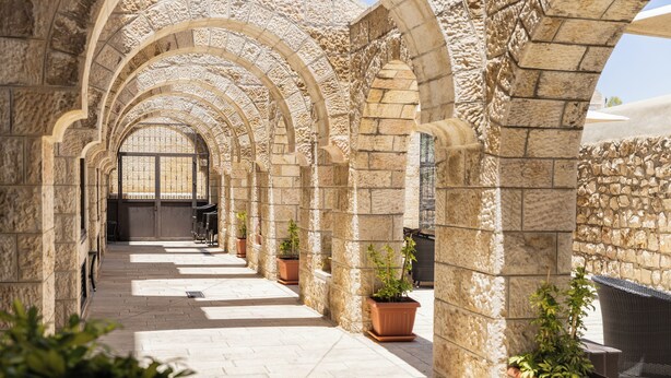 A corridor with stone arches, potted plants, and a view of a building in the distance. - New Gate Hostel (Jerusalem)