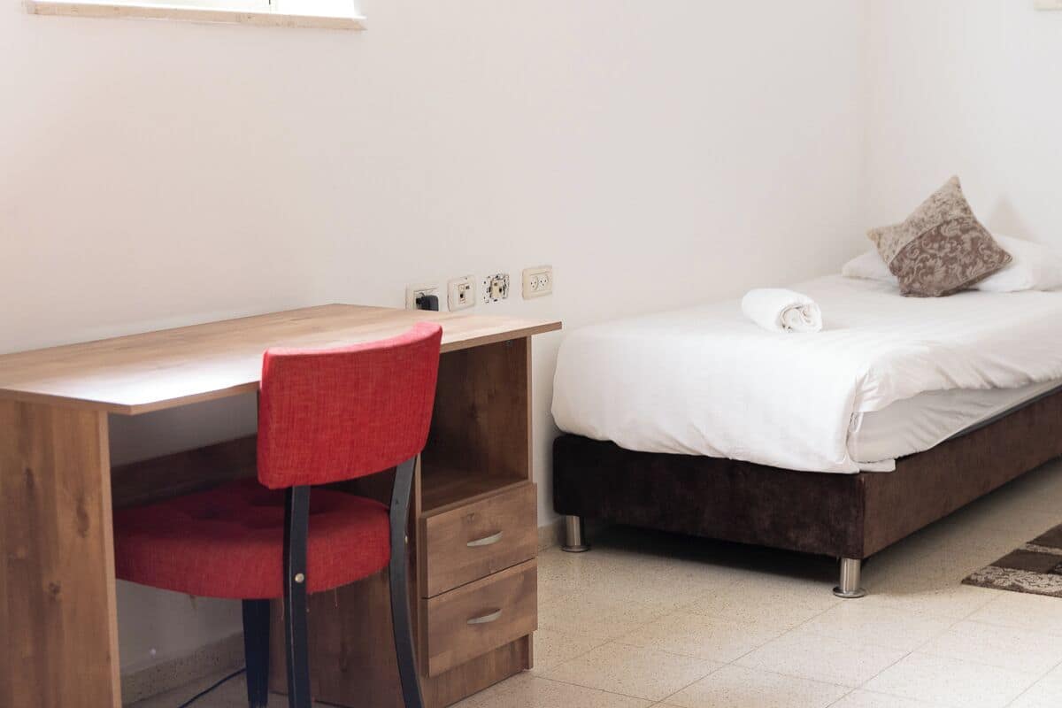 A bedroom with a wooden desk, a red chair, a bed with white bedding, and a patterned pillow.