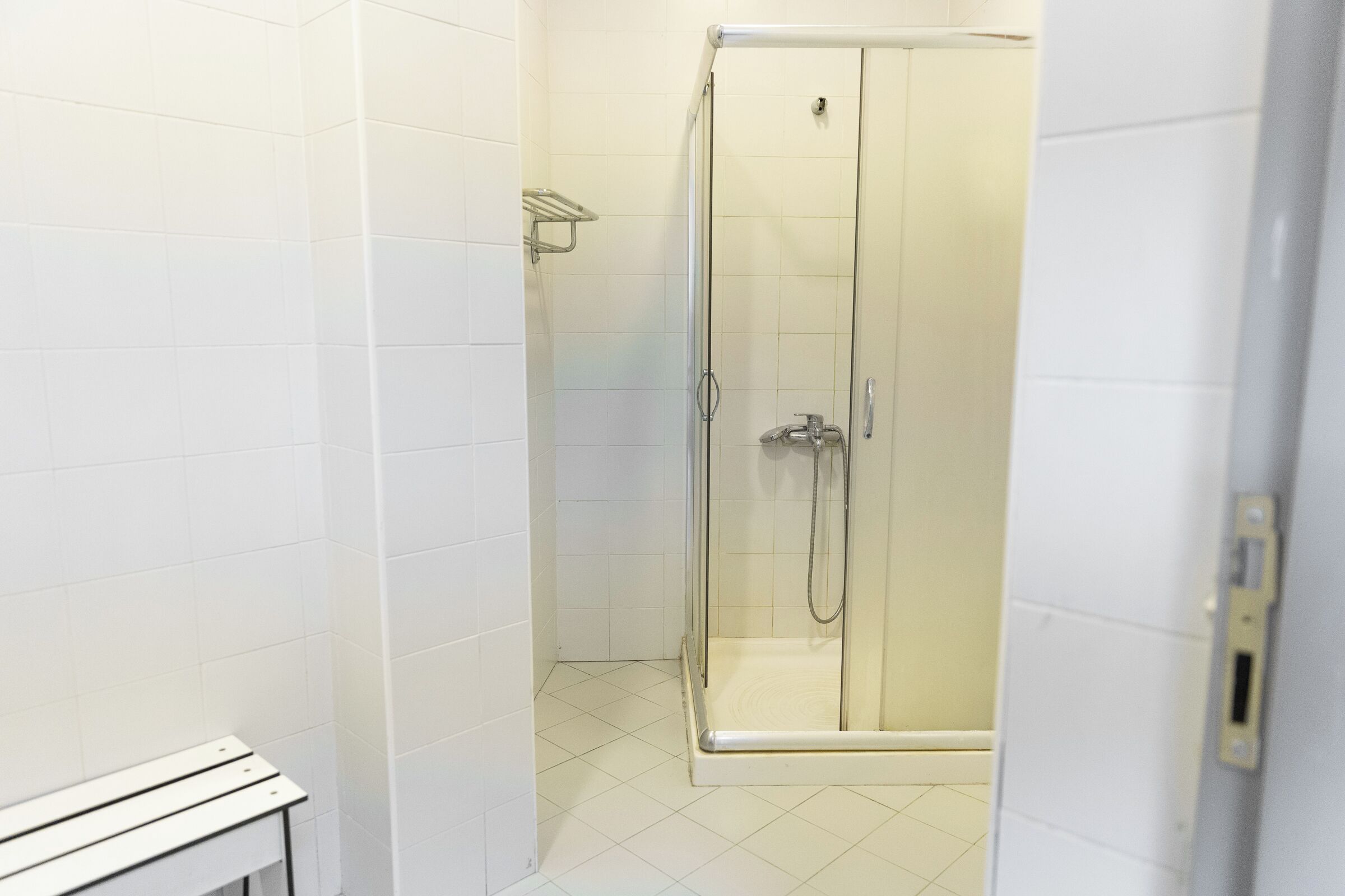 Une salle de bain moderne avec des murs et un sol recouverts de carreaux blancs, une douche à parois de verre et un banc blanc.