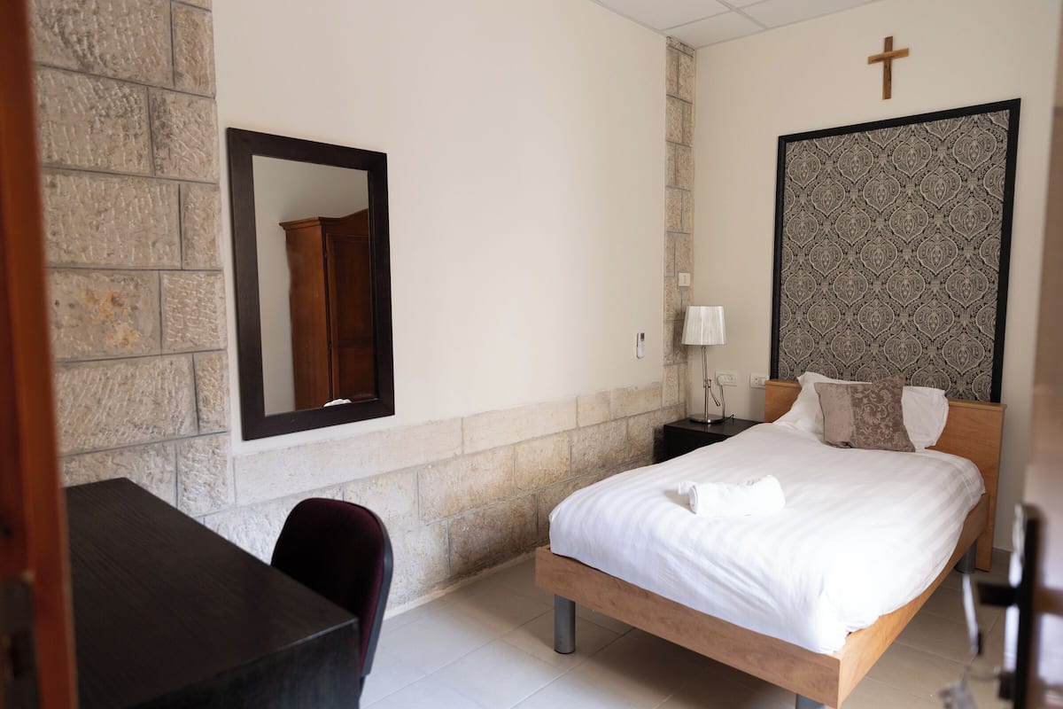 A bedroom with a bed, a desk, a chair, a nightstand, a lamp, and a cross on the wall.