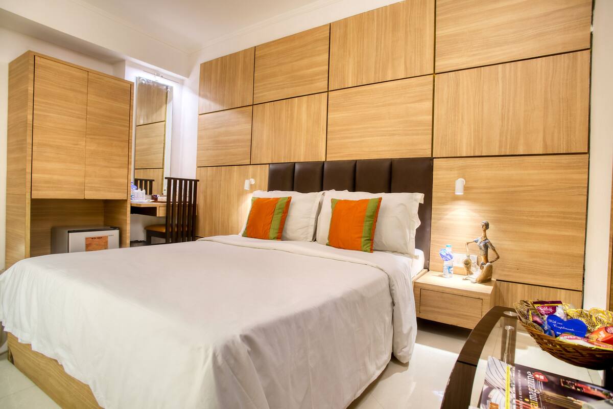 Una habitación de hotel con una cama grande, paredes de paneles de madera, una mesita de noche con una lámpara y una pequeña mesa con una cesta de objetos.