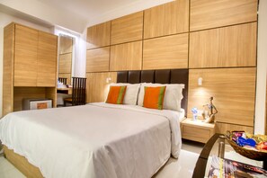 A hotel room with a large bed, wooden panel walls, a nightstand with a lamp, and a small table with a basket of items.