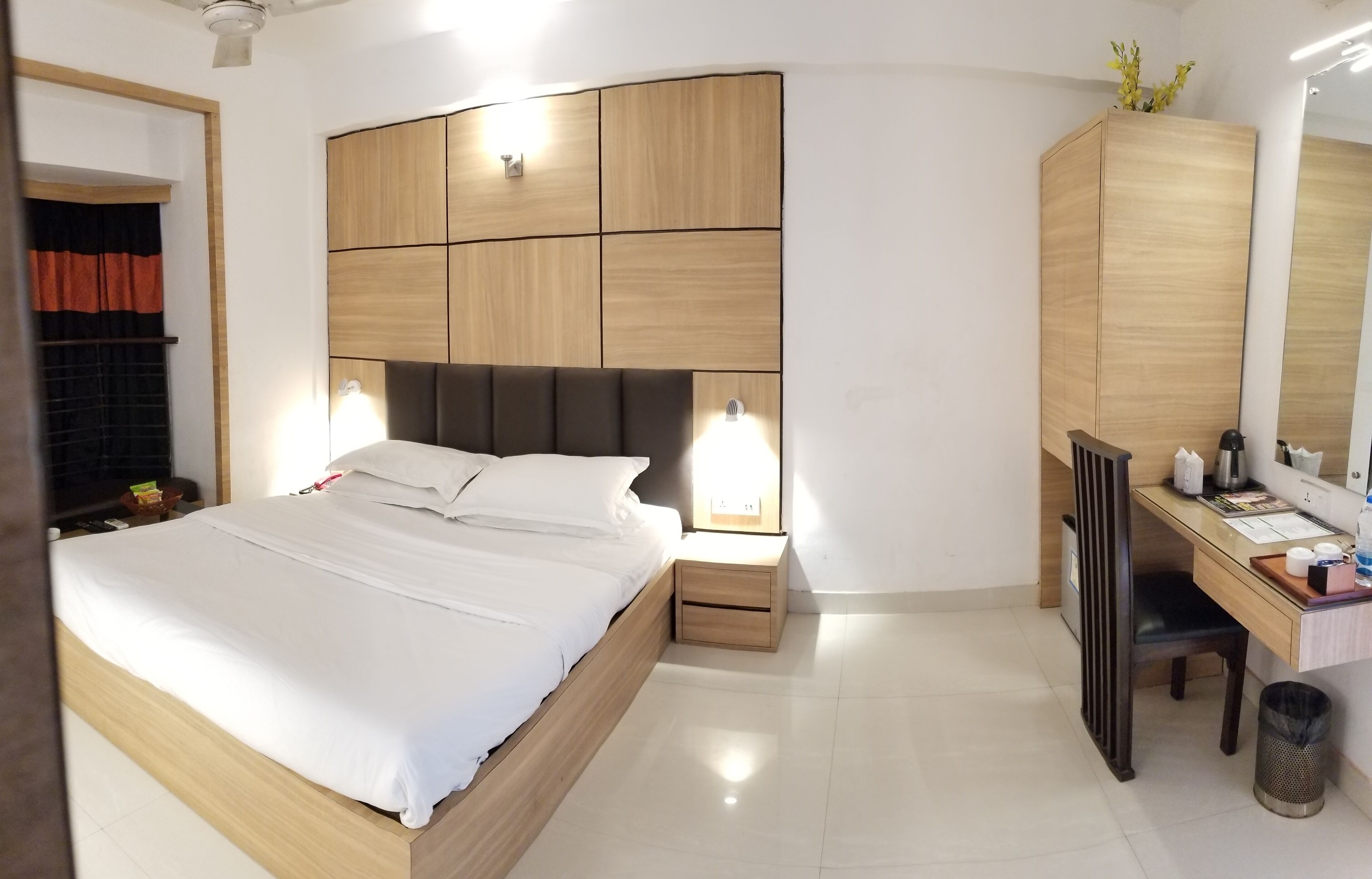 A modern bedroom with a wooden headboard, a bed with white linens, a desk with a chair, and a mirror.