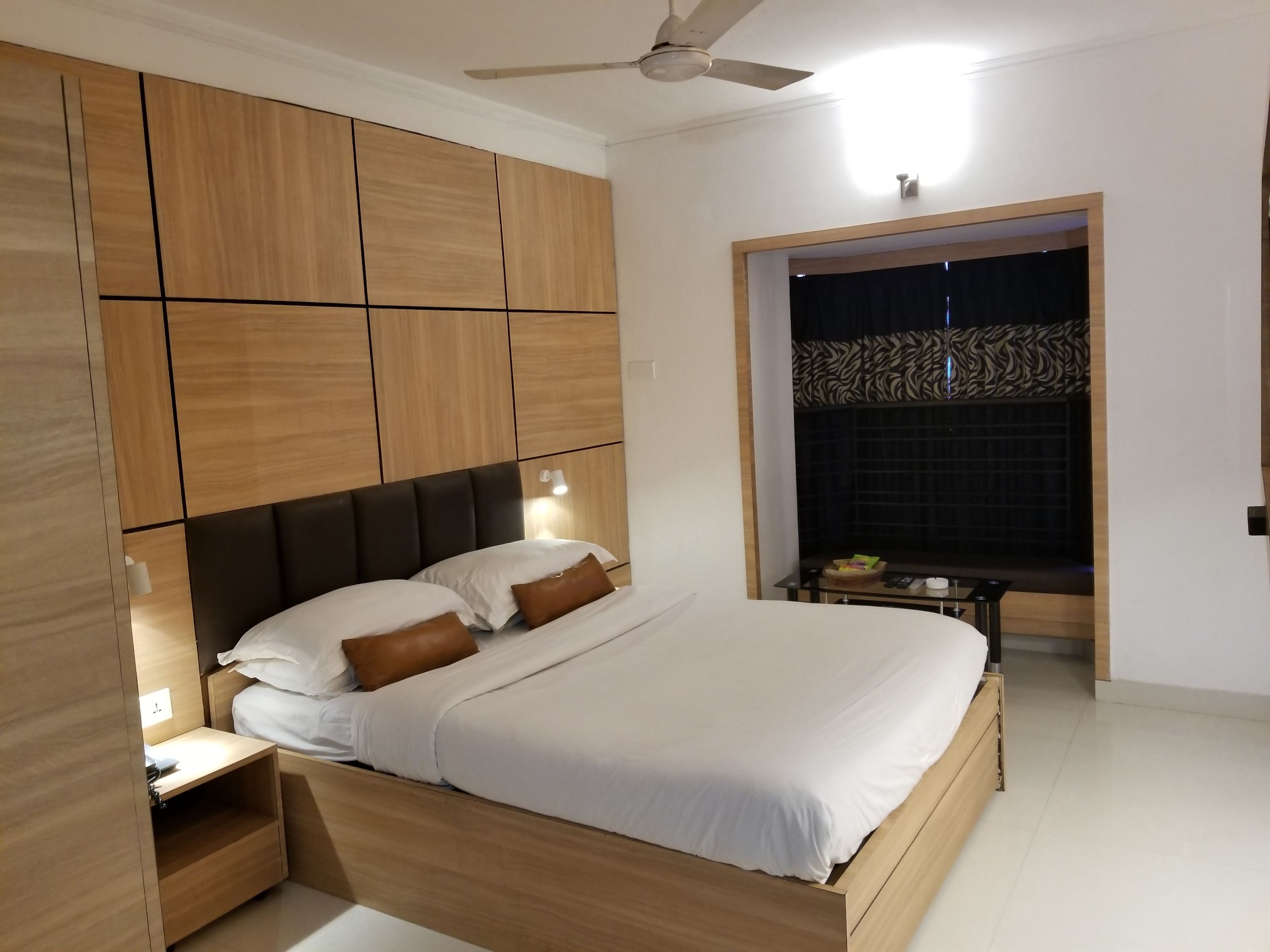 Un dormitorio moderno con cabecera de madera, cama con ropa blanca, mesita de noche y ventilador de techo.