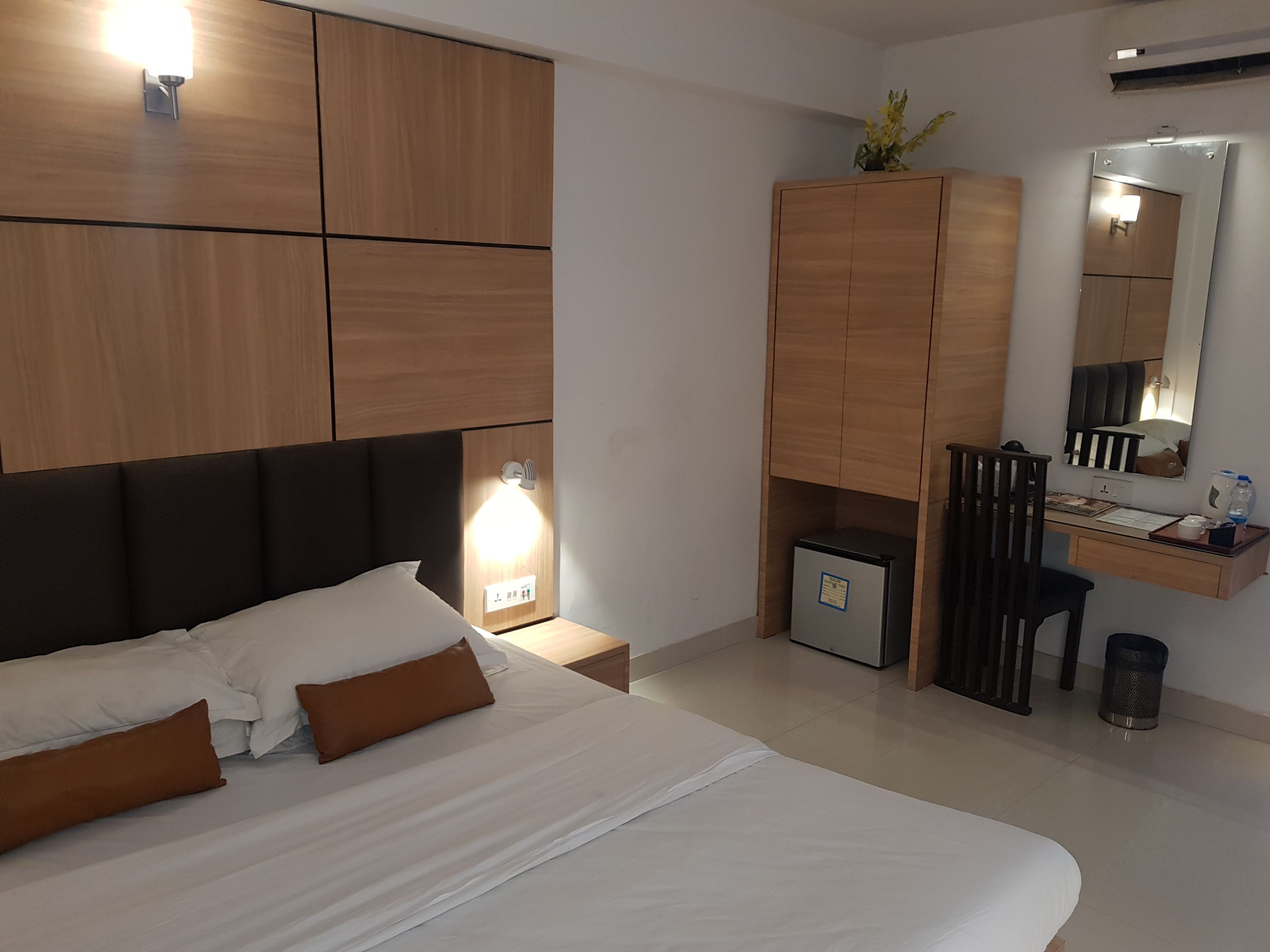 A modern hotel room with a large bed, a desk with a computer, a small fridge, and a mirror.