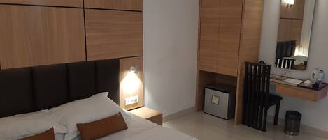 A modern hotel room with a large bed, a desk with a computer, a small fridge, and a mirror.