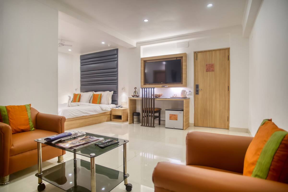 Una habitación de hotel moderna con una cama, dos sofás, un escritorio con computadora, un televisor y un refrigerador pequeño.