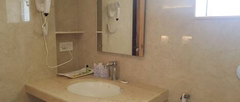 Un baño con un lavabo de mármol color crema, un espejo, un secador de pelo, un perchero para toallas y un dispensador de papel higiénico.