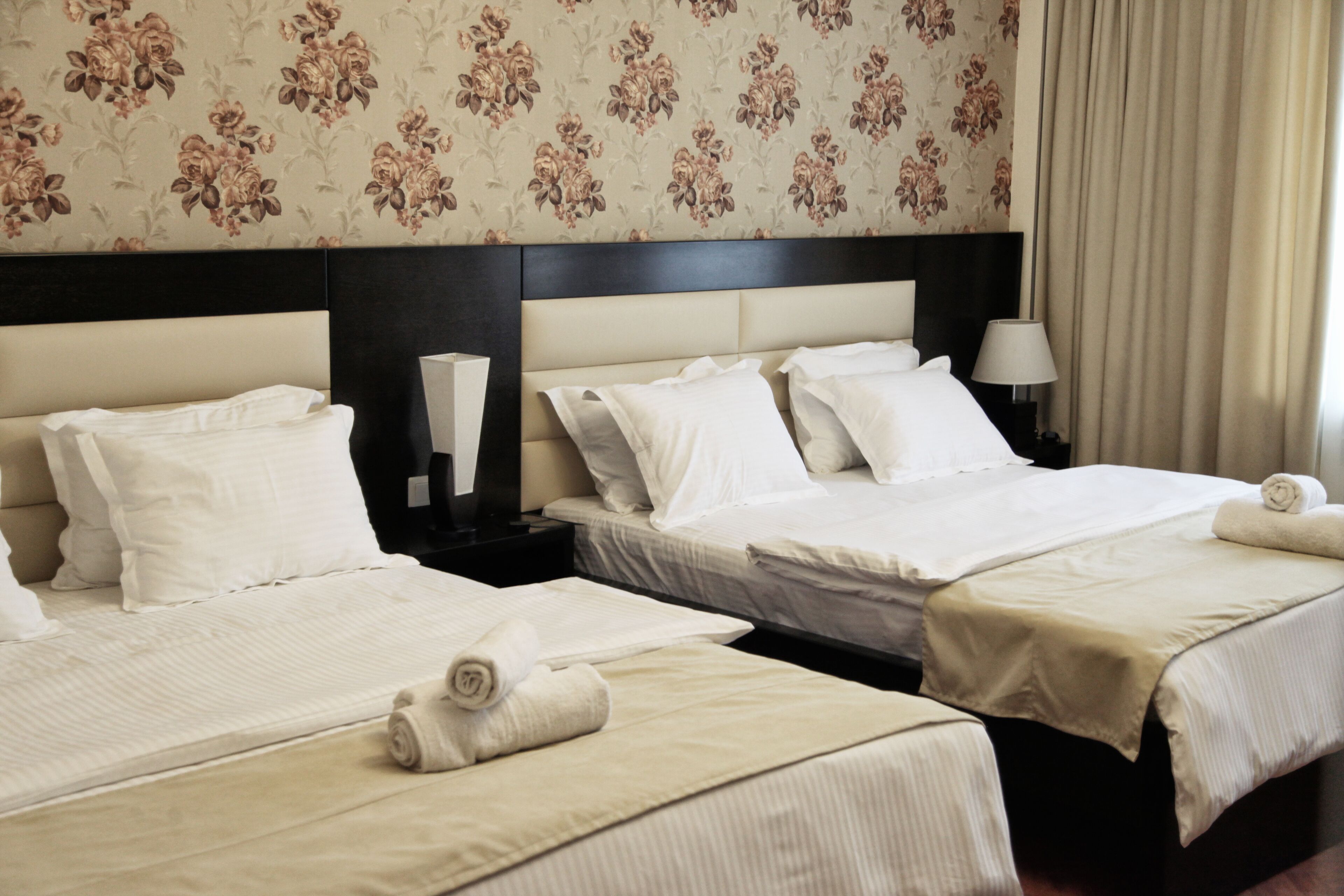 A hotel room with two beds, each with white linens and pillows, a headboard, bedside lamps, and a floral wallpaper pattern.