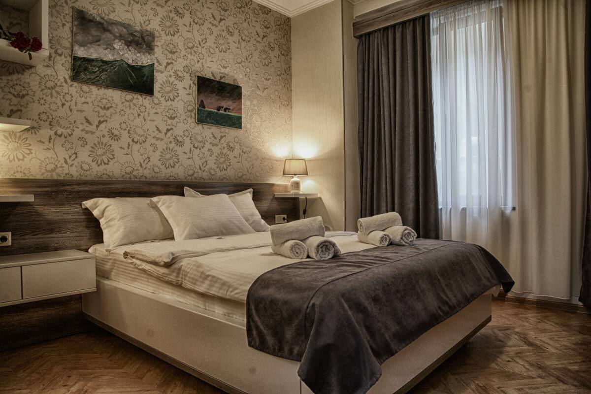 A modern bedroom with a large bed, a nightstand, a lamp, and a wall with floral wallpaper and two paintings.