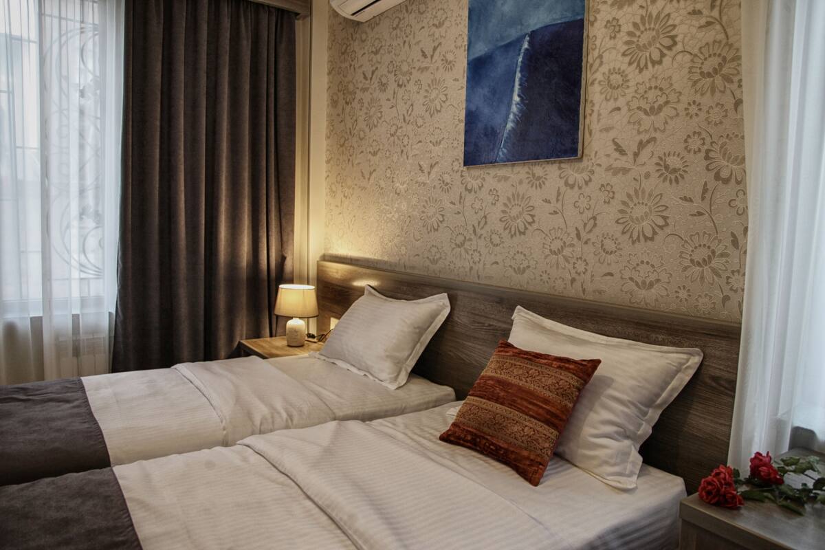 A hotel room with two beds, a nightstand with a lamp, floral wallpaper, and a painting on the wall.