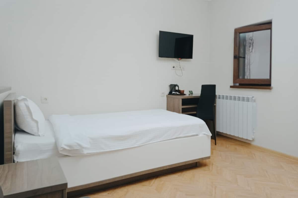 A bedroom with a bed, a desk, a chair, a television mounted on the wall, and a window with a mirror.