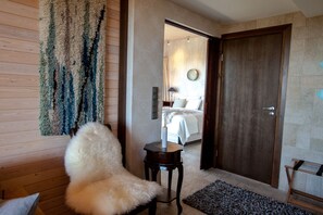 A hotel room with a wooden floor, a fluffy chair, a small table, a bed with a white bedspread, a wooden door, and a wall-mounted mirror.