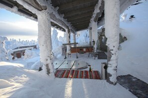 A snow-covered outdoor area with a wooden bench and a table, surrounded by snow-covered trees and a stone wall.