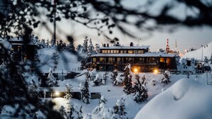 A snow-covered building with multiple windows and a lit interior, surrounded by snow-laden trees and a ski slope in the background. - Ruka Peak (Kuusamo)