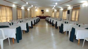 A conference room with rows of tables and chairs, a stage at the front, and ceiling lights. - Kirigime Guest House (Kabale)