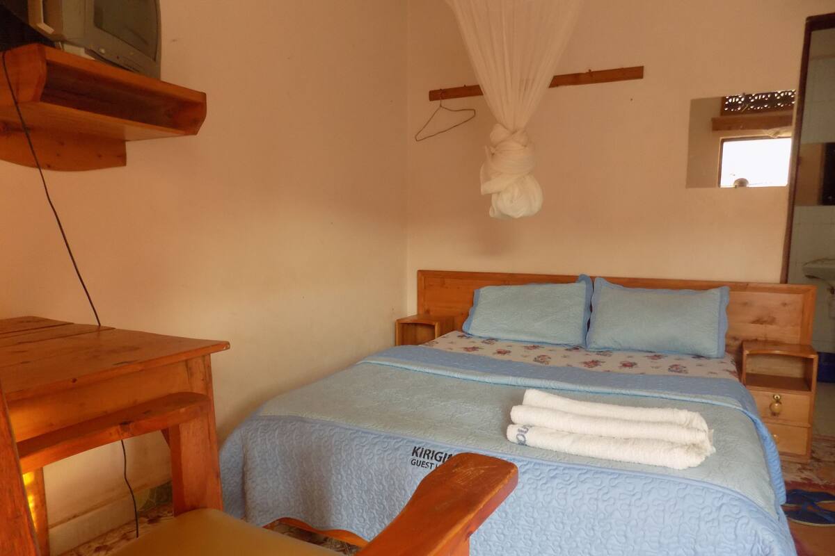 A bedroom with a wooden bed, a bedside table, a chair, a hanging mosquito net, and a television on a wooden shelf.