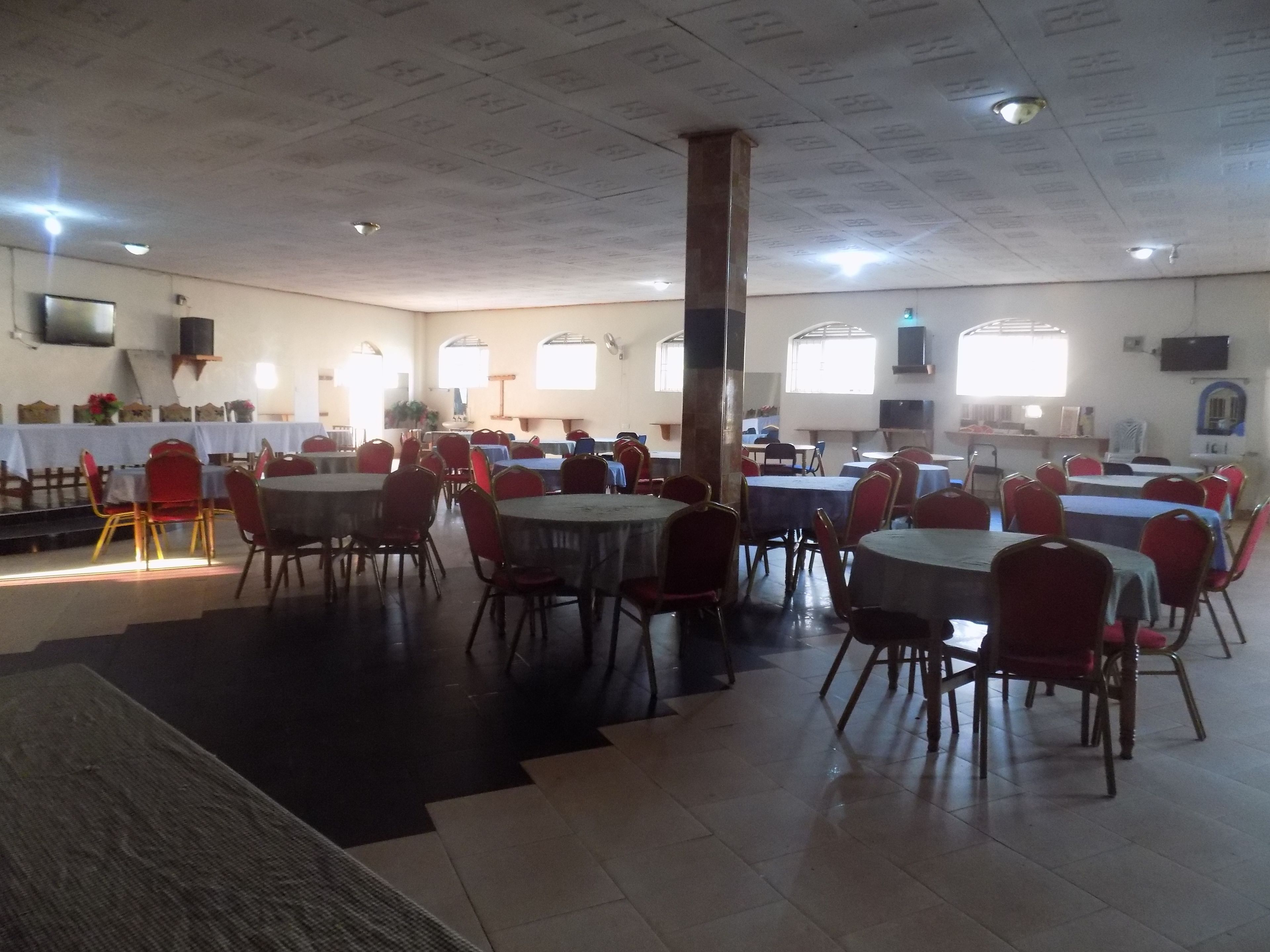 A spacious dining hall with multiple round tables and chairs, a high ceiling with recessed lighting, and a stage area in the background.