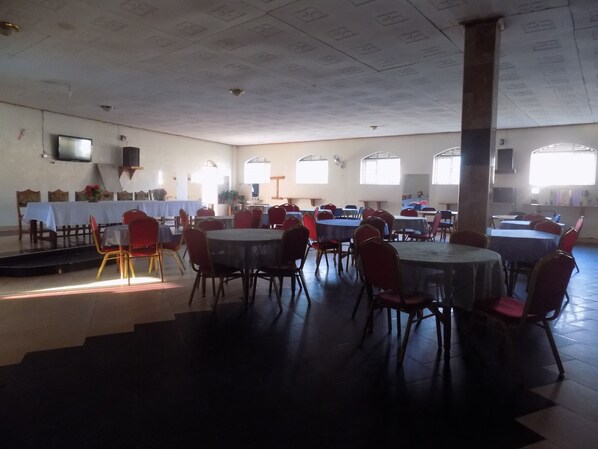 Un salón amplio con varias mesas redondas y sillas, un escenario al frente y grandes ventanales que dejan entrar luz natural.