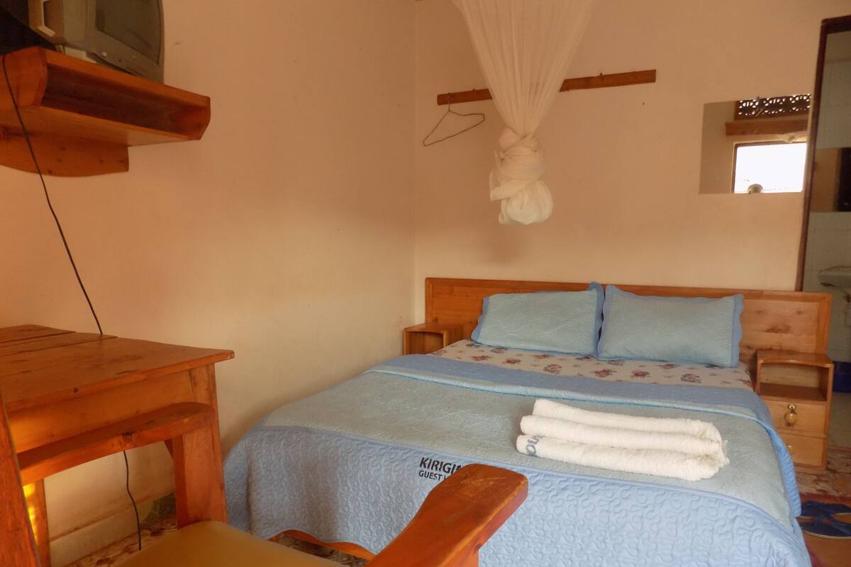 A bedroom with a bed, a wooden nightstand, a bedside table, a hanging white canopy, and a television on a wooden shelf.