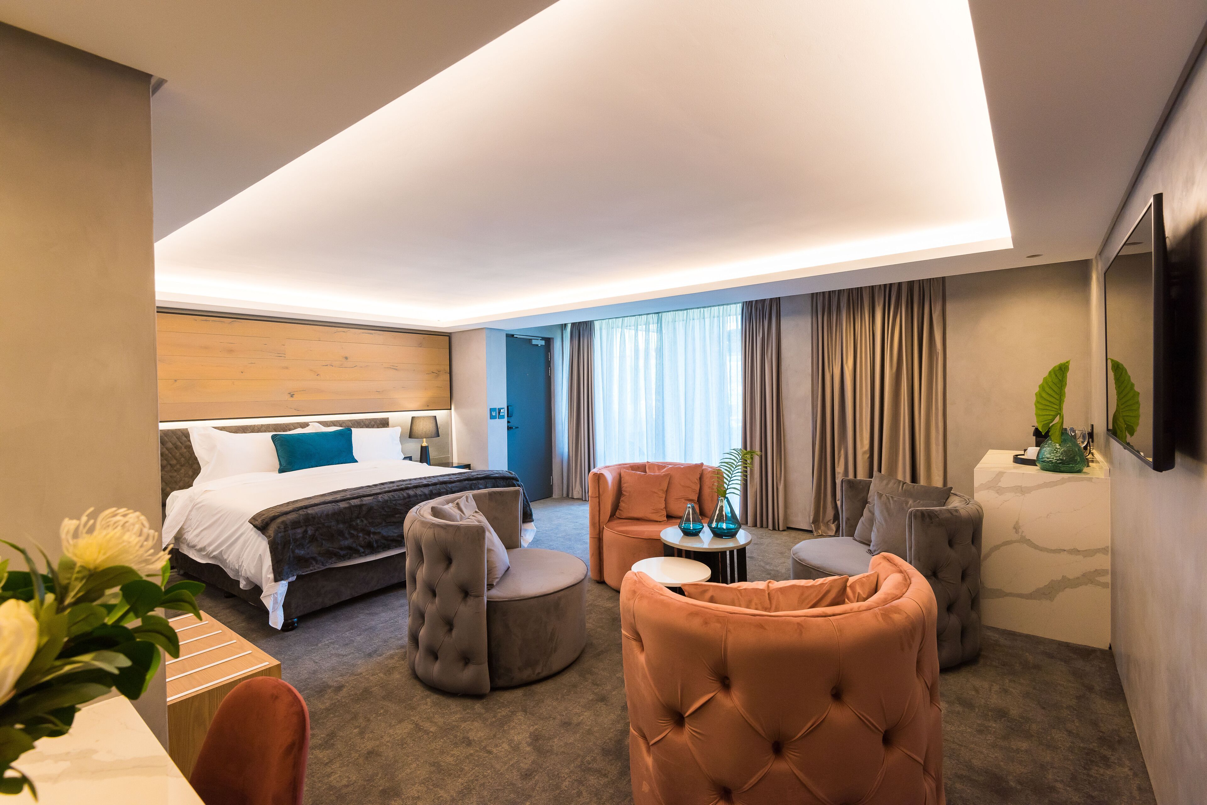 A modern hotel room with a bed, two ottomans, a sofa, a small table, and a TV mounted on the wall.